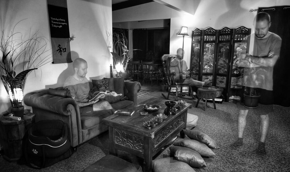 Man pictured on couch, in chair and standing with arms crossed in black and white photo