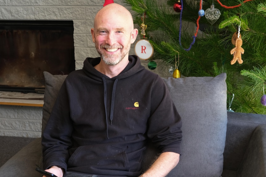 A bald man smiles at the camera. There's a Christmas Tree behind him.