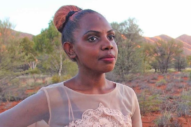 Indigenous teenage girl with hair pulled back in tight bun, posing in desert landscape wearing pink formal dress