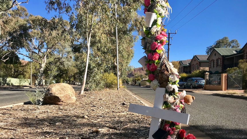 A white cross stands next to a tree with pink and white flowers, fairy lights and a teddy bear stuck to the trunk