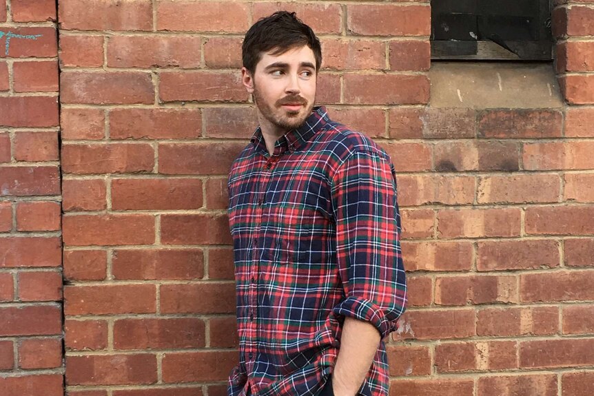 A brown haired man in a plaid shirt leans against an exposed brick wall, looking away from the camera