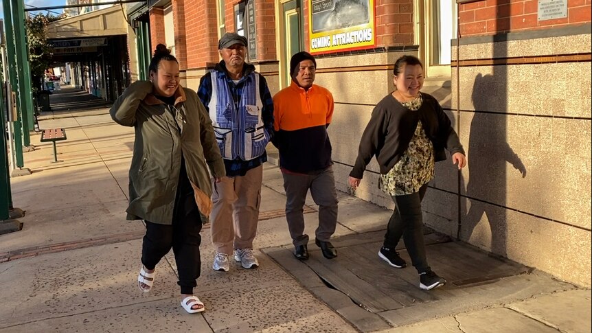 Four people of south-east Asian descent walk down a street with a regional pub behind. They are smiling.