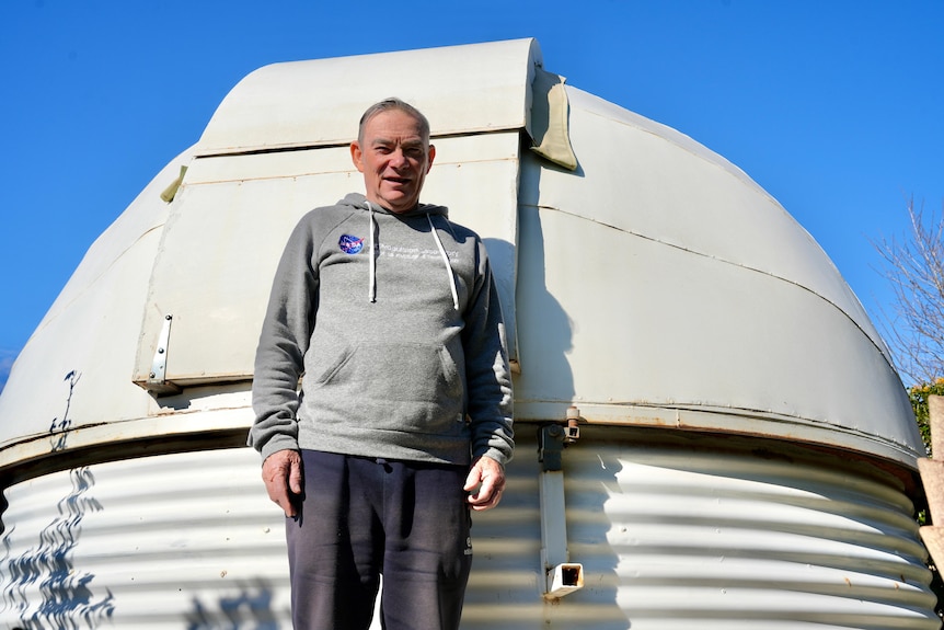 A man stands smiling next to a white domed observatory with a clear blue sky in the background.