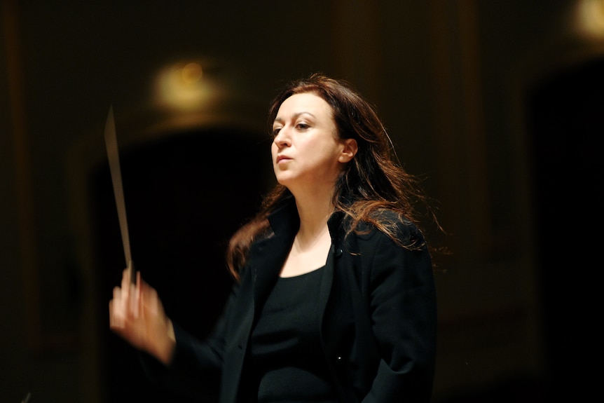 White middle-aged woman with long brown hair wears all black and stands on a podium wielding a conductor's baton.