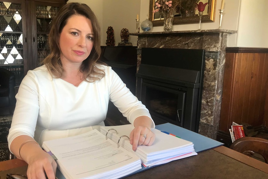 Ms Tranter sits with a large binder of pages. A large bookcase filled with what look like legal texts behind her.