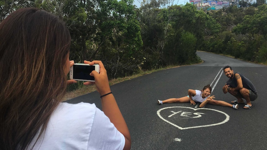 Three Japanese tourists take photos, one of them does the leg splits in the middle of the road, the other sits.