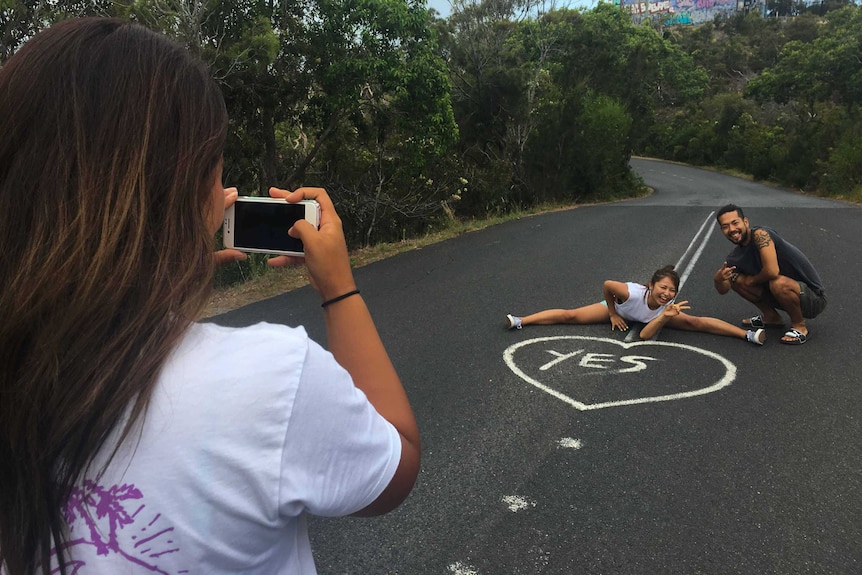 Three Japanese tourists take photos, one of them does the leg splits in the middle of the road, the other sits.