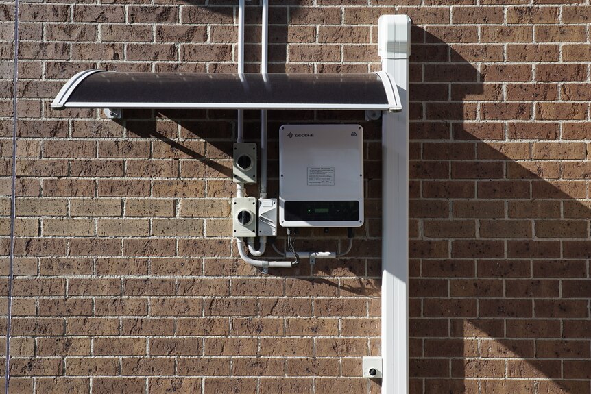 A new solar panel inverter, which looks like a white plastic box connected to pipes leading to the roof, on a brick wall.