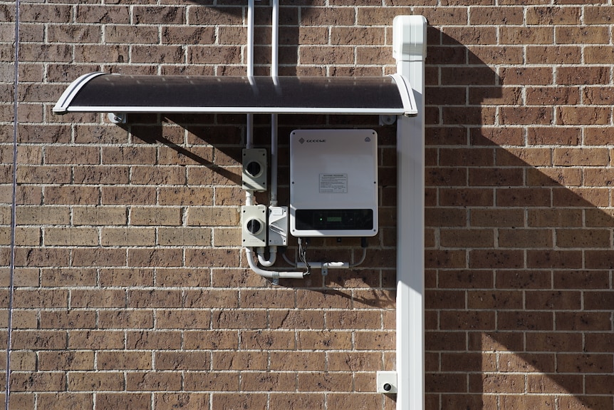 A new solar panel inverter, which looks like a white plastic box connected to pipes leading to the roof, on a brick wall.