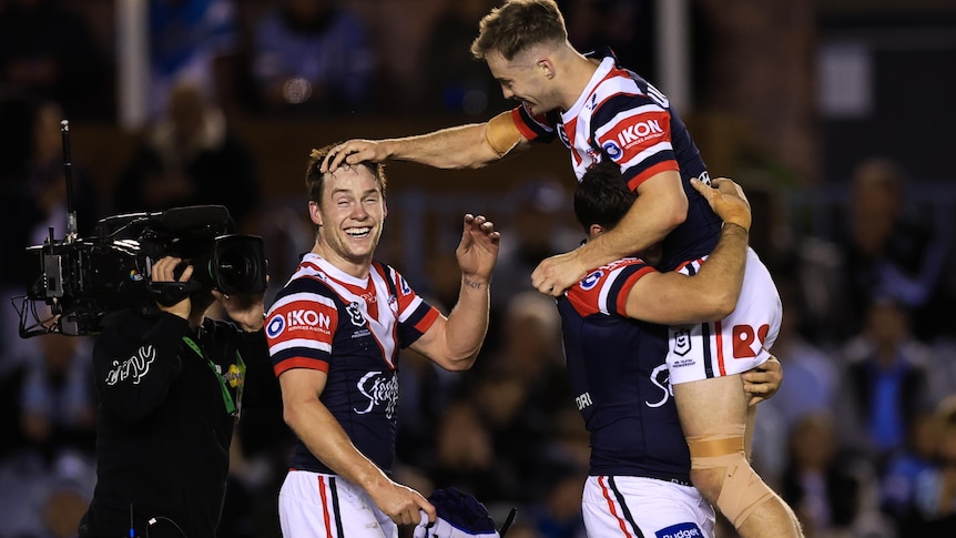Three male rugby league players embrace after winning a game of football, wearing red, white and blue shirts