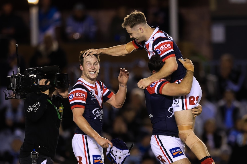 Three male rugby league players embrace after winning a game of football, wearing red, white and blue shirts
