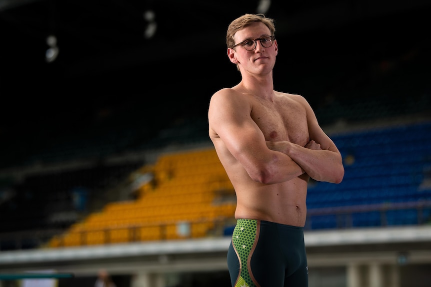 Australian Olympic swimmer Mack Horton stands next to a pool in swim gear with his arms folded. 