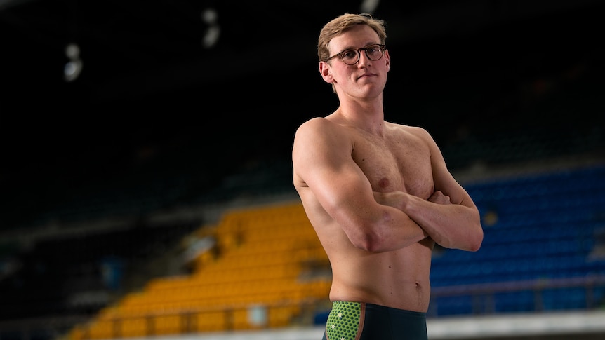 Australian Olympic swimmer Mack Horton stands next to a pool in swim gear with his arms folded. 