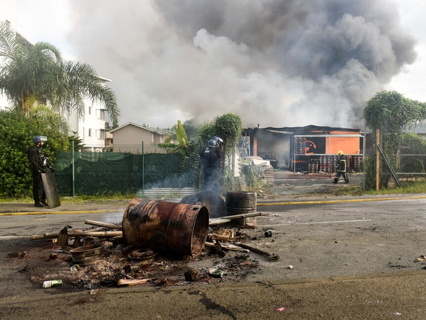 A burnt-out barrel and debris sits on a road in front of a burning building which a fire fighter is walking toward