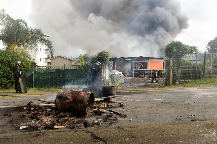 A burnt-out barrel and debris sits on a road in front of a burning building which a fire fighter is walking toward