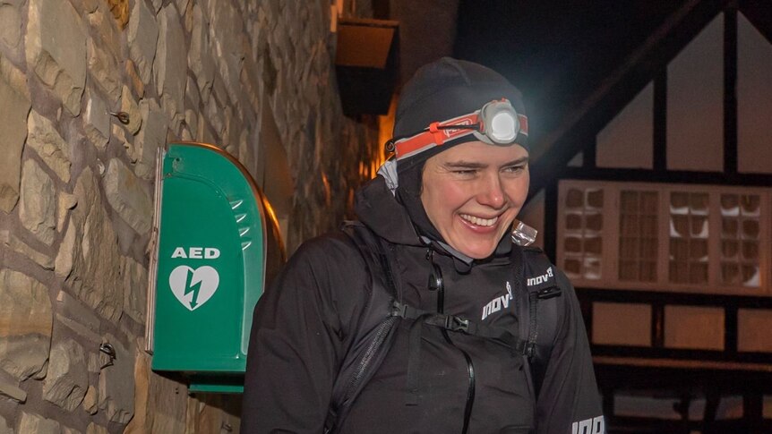 A woman in a black jacket wearing a headlamp, smiling against a cobblestone wall.