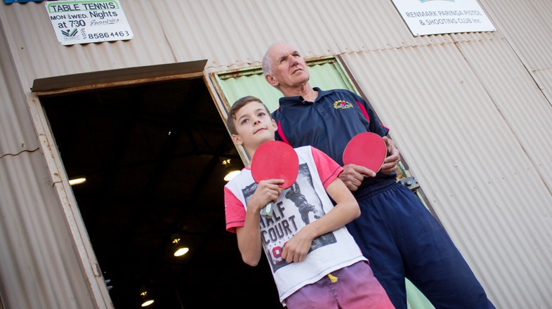 Evan McAllister, 10 and veteran Dennis Robinson hold red table tennis bats and stand outside shed below table tennis sign.