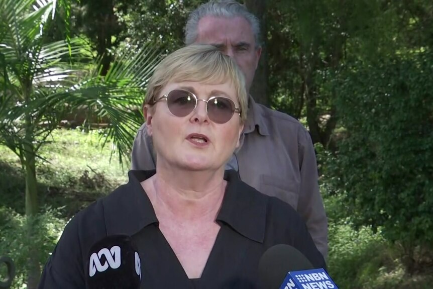 A middle-aged woman wearing glasses speaking to a group of journalists 