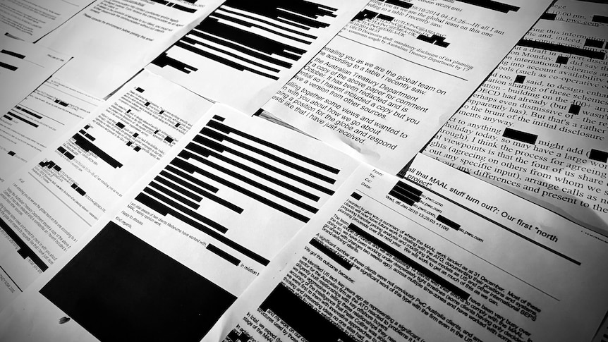 Printed black and white emails lay on a table with large black boxes redacting text.