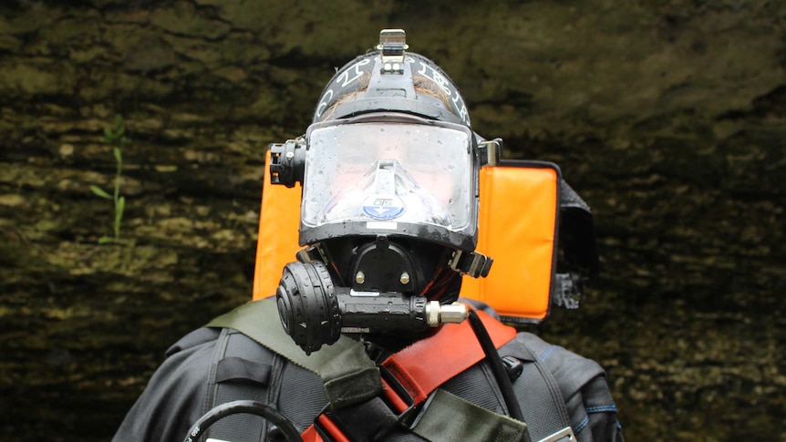 a man wearing a full diving suit emerges up steps from a cave