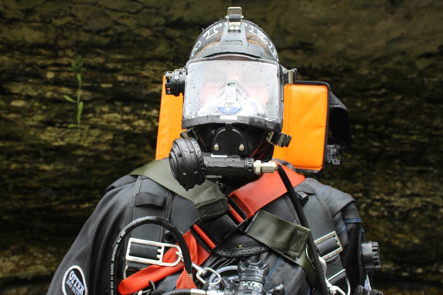 a man wearing a full diving suit emerges up steps from a cave