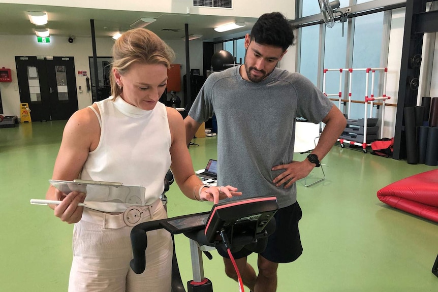 Suzy Russell, stands beside a male athlete, entering data on an exercise bike.