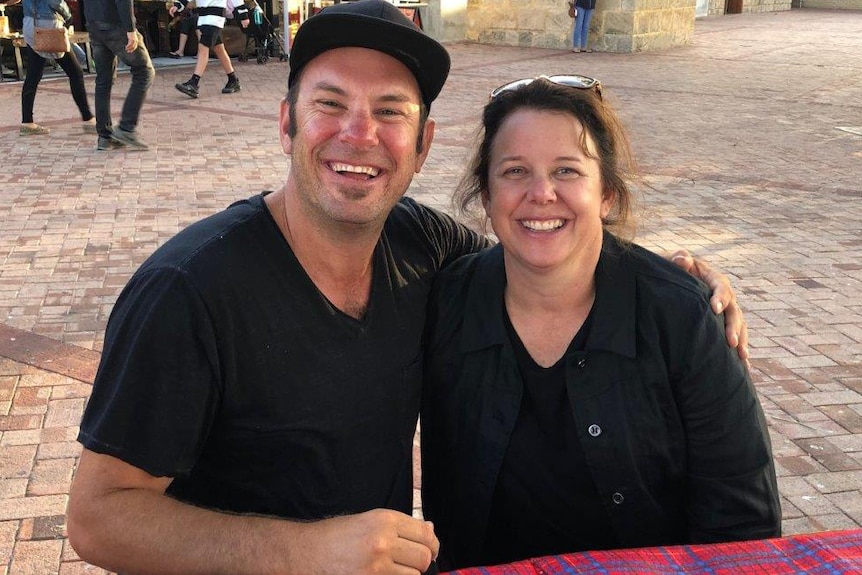 A middle-aged man and woman sit next to each other smiling for the camera on a bench in a public courtyard.