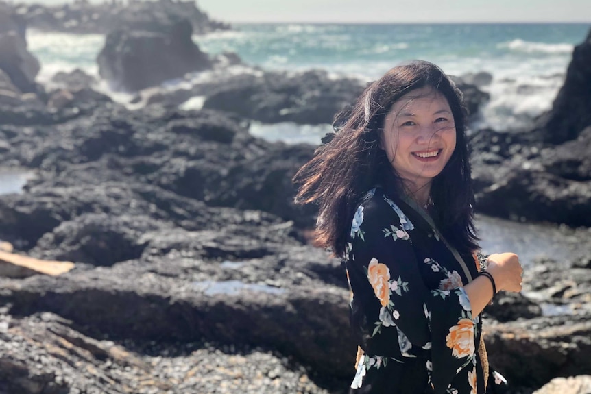 A woman with dark hair smiling towards the camera, with waves crashing towards the rocks behind her.