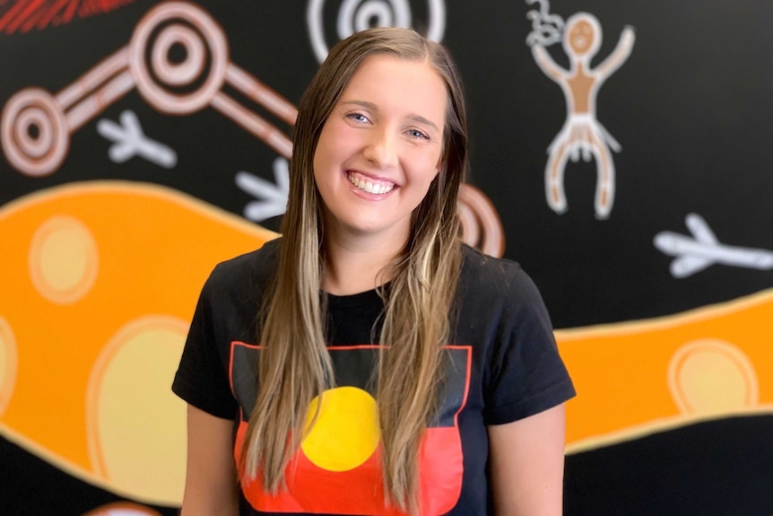 A woman in a shirt with an Aboriginal Flag smiles at the camera, with indigenous artwork behind her