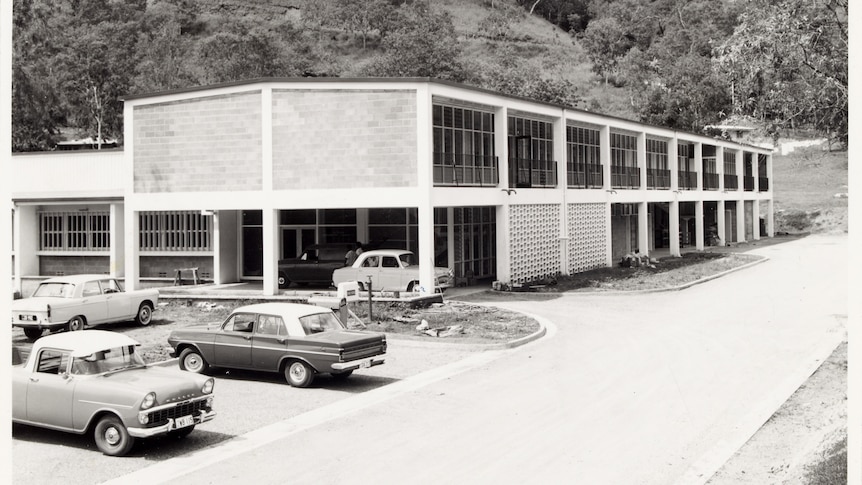 Double storey section of a building set at the foot of a hill. Some cars of the era in the carpark. Landscaping not finished.