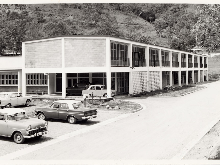 Double storey section of a building set at the foot of a hill. Some cars of the era in the carpark. Landscaping not finished.