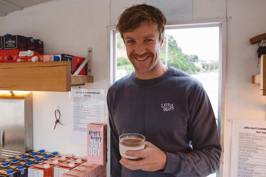 A man with short hair and a moustache holds and takeaway coffee and smiles for the camera