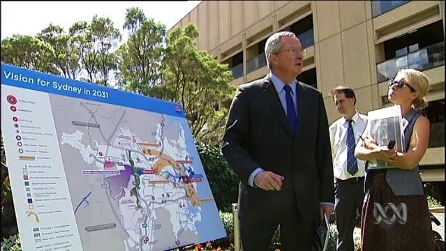 Politician stands beside large poster at press conference, poster shows map of Sydney and reads "Vision for Sydney in 2031"
