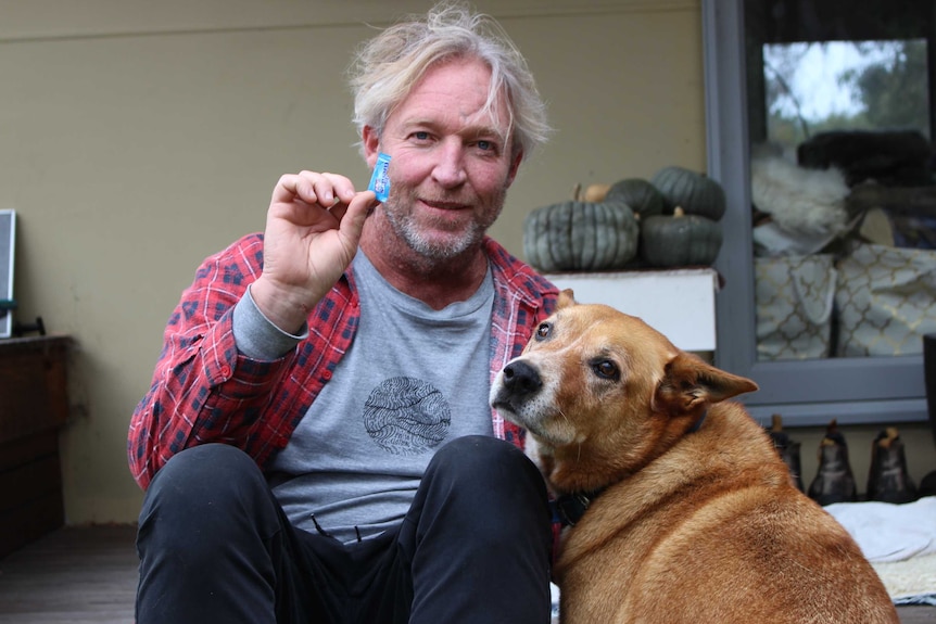 A middle-aged man sits on his verandah with his dog and holds a tiny piece of blue plastic.