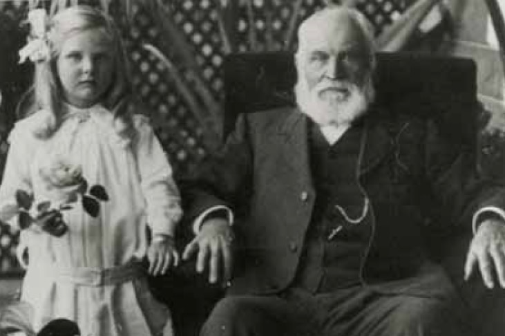 Black and white image of an old man with a white beard dressed in a suit sitting in a large chair next to a standing young girl.