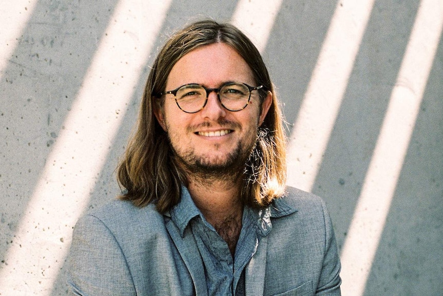 Cooney, wearing long hair and horn-rimmed spectacles, stands in dappled light against a cement wall.