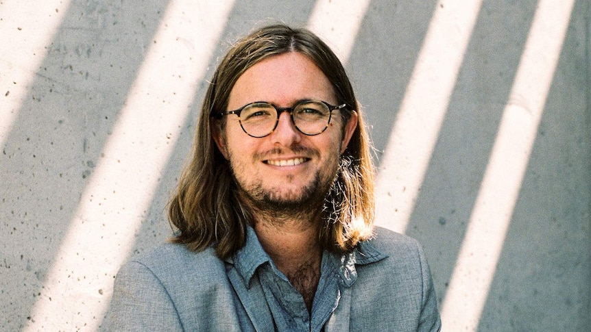 Cooney, wearing long hair and horn-rimmed spectacles, stands in dappled light against a cement wall.