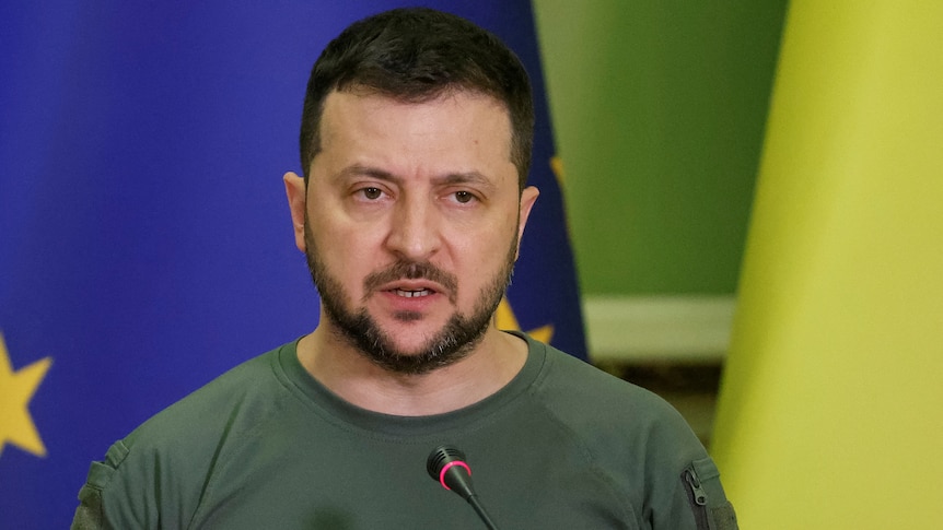 A man in a khaki T shirt speaks into a thin microphone while standing in front of EU and Ukraine flags.