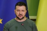 A man in a khaki T shirt speaks into a thin microphone while standing in front of EU and Ukraine flags.