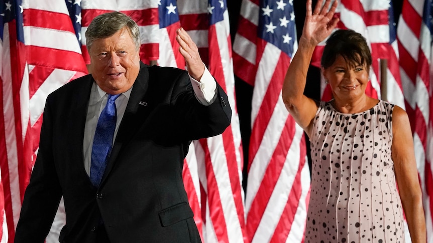 Viktor Knavs and Amalija Trump wave to a crowd in front of american flags. She wears a spotty dress with hair up