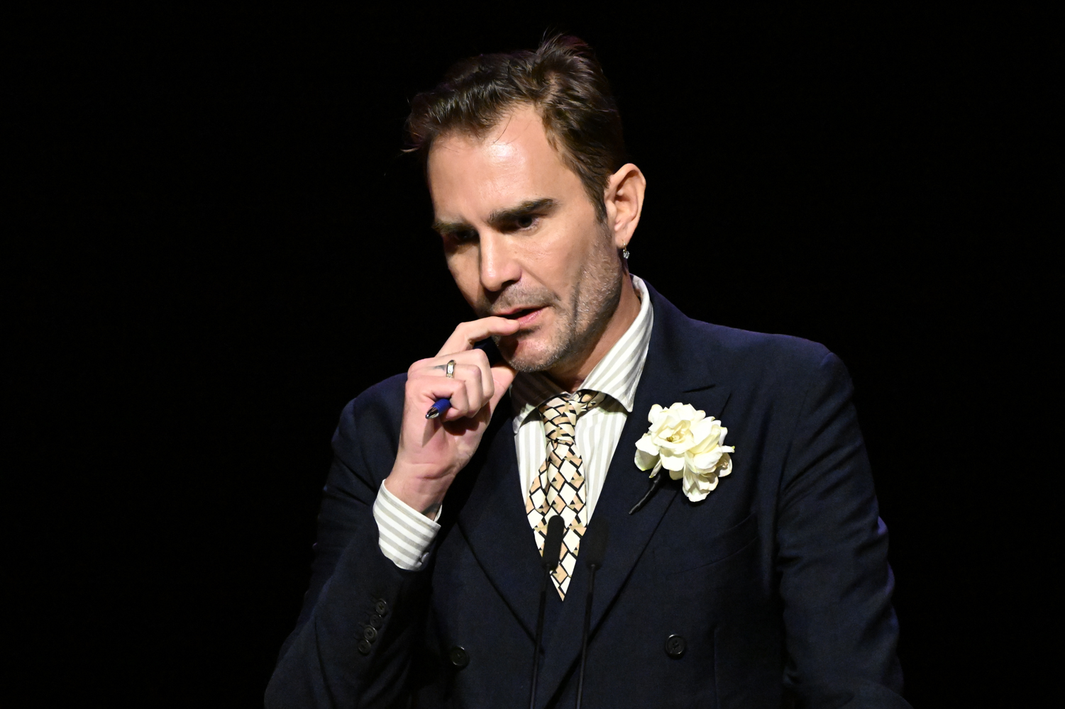 Oscar Humphries holds his had up to his face in thought while a white flower is pinned to his jacket