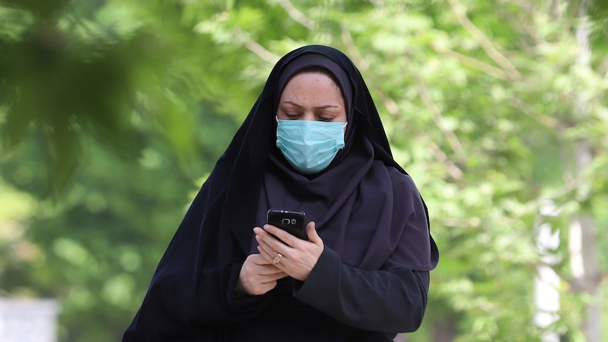 A woman wearing a hijab and surgical-style mask looks at a mobile phone. 