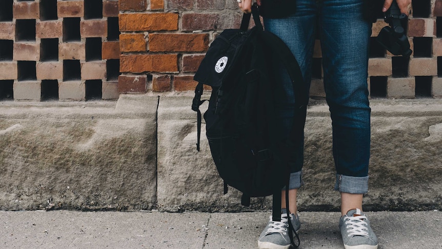 A student stands against a brick wall holding a backpack. You can only see their feet and legs and the bag.