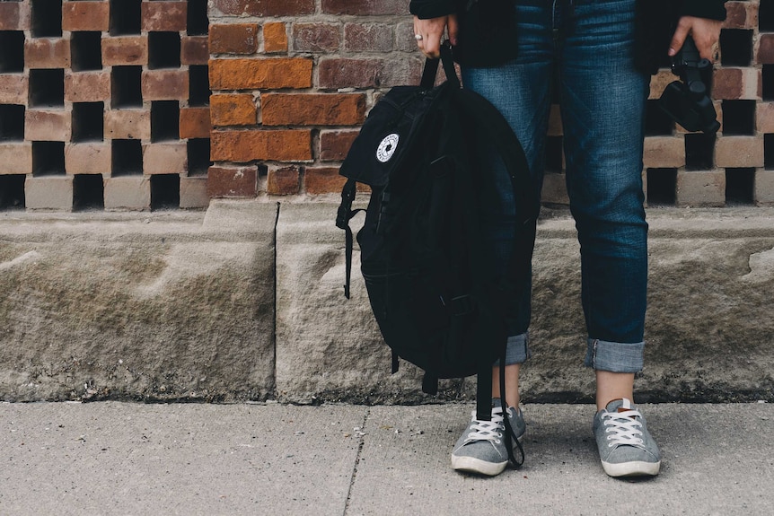 A student stands against a brick wall holding a backpack. You can only see their feet and legs and the bag.