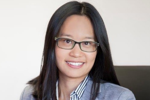 A woman in glasses sits at a desk and smiles