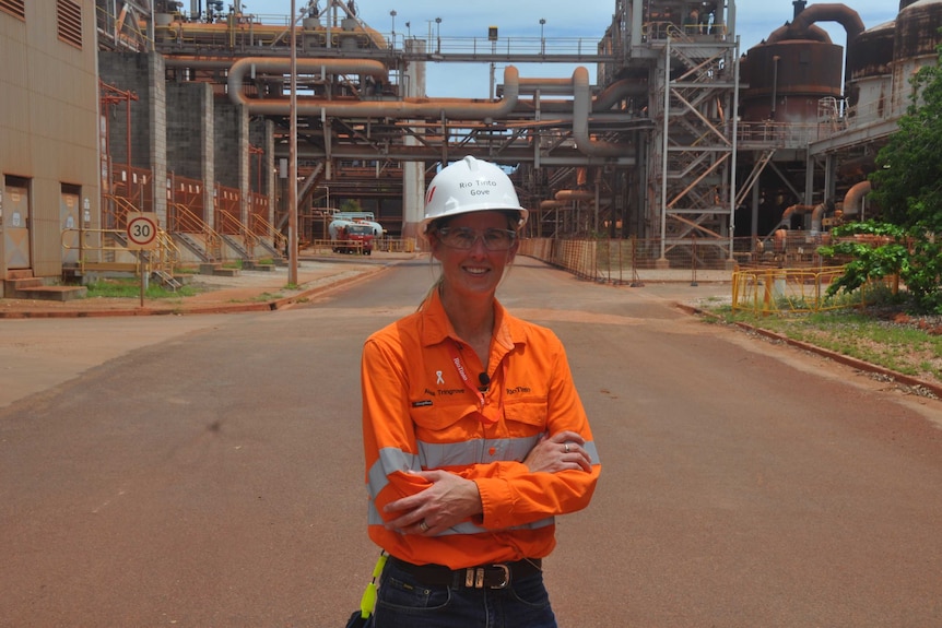 Alysia Tringrove is wearing an orange work shirt and a white hard hat. Her arms are folded and she smiles at the camera.