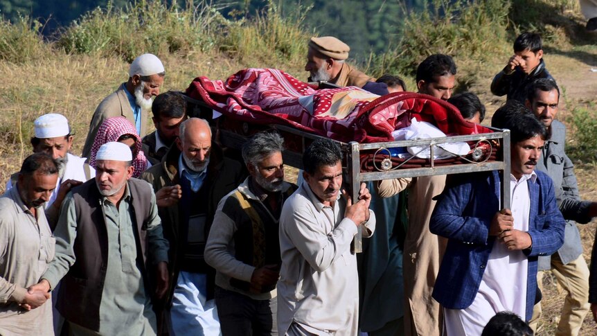Several men carry the body of a man wrapped in a red blanket to their burial