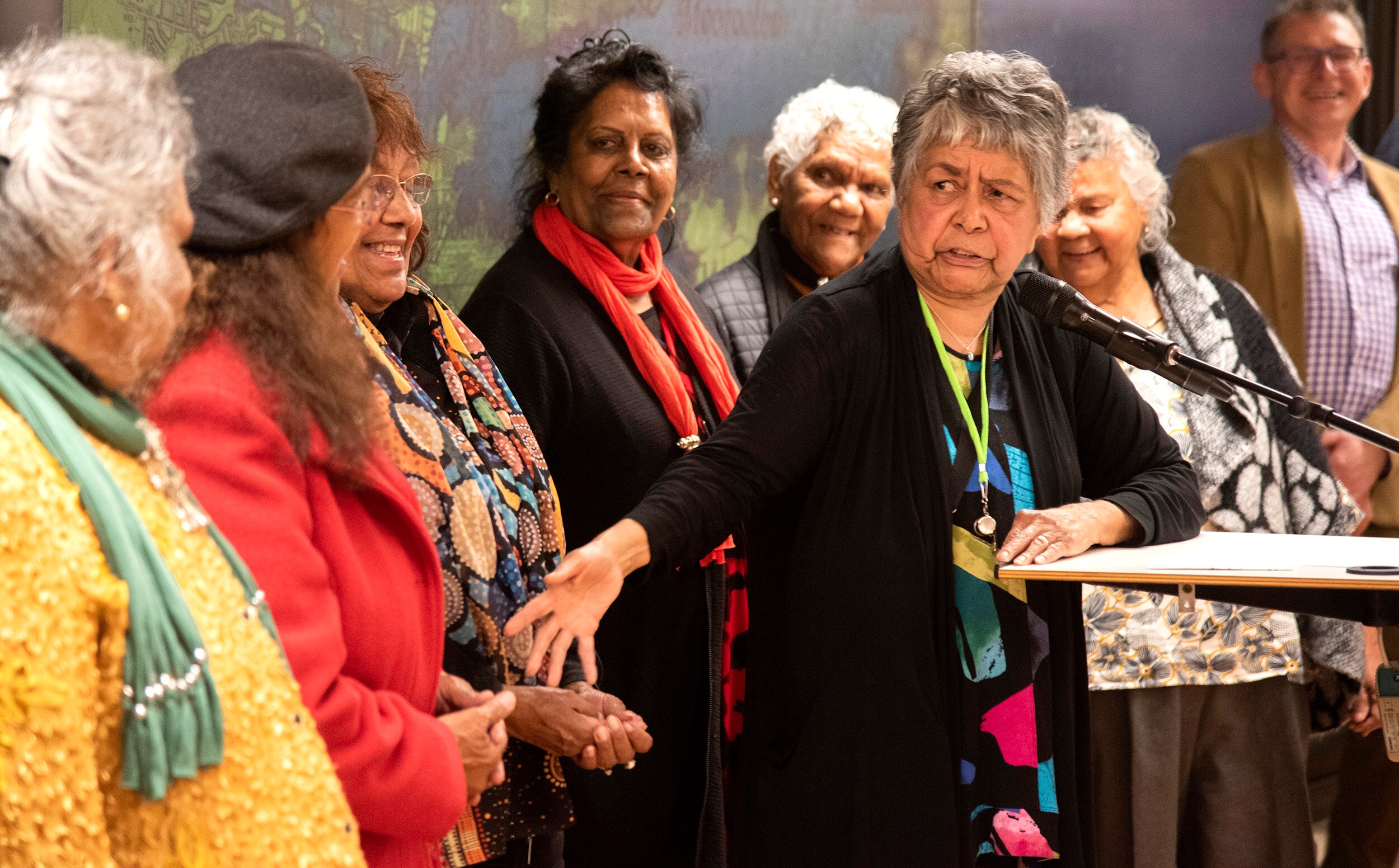 A group of women stand behind a woman speaking at a podium.