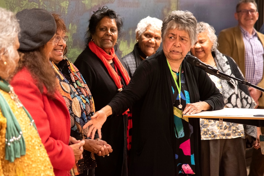 A group of women stand behind a woman speaking at a podium.