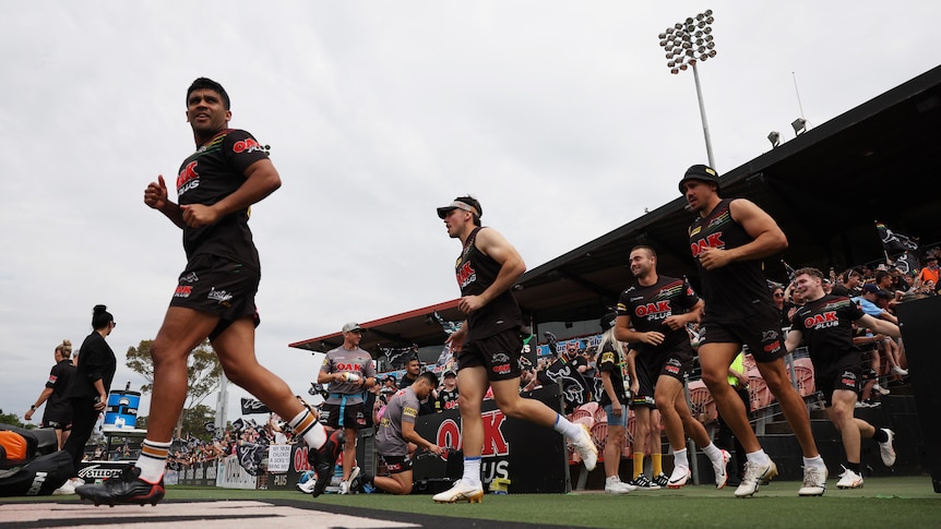 A Penrith Panthers NRLW player runs out on the ground in training gear, followed by teammates as a big crowd watches on. 